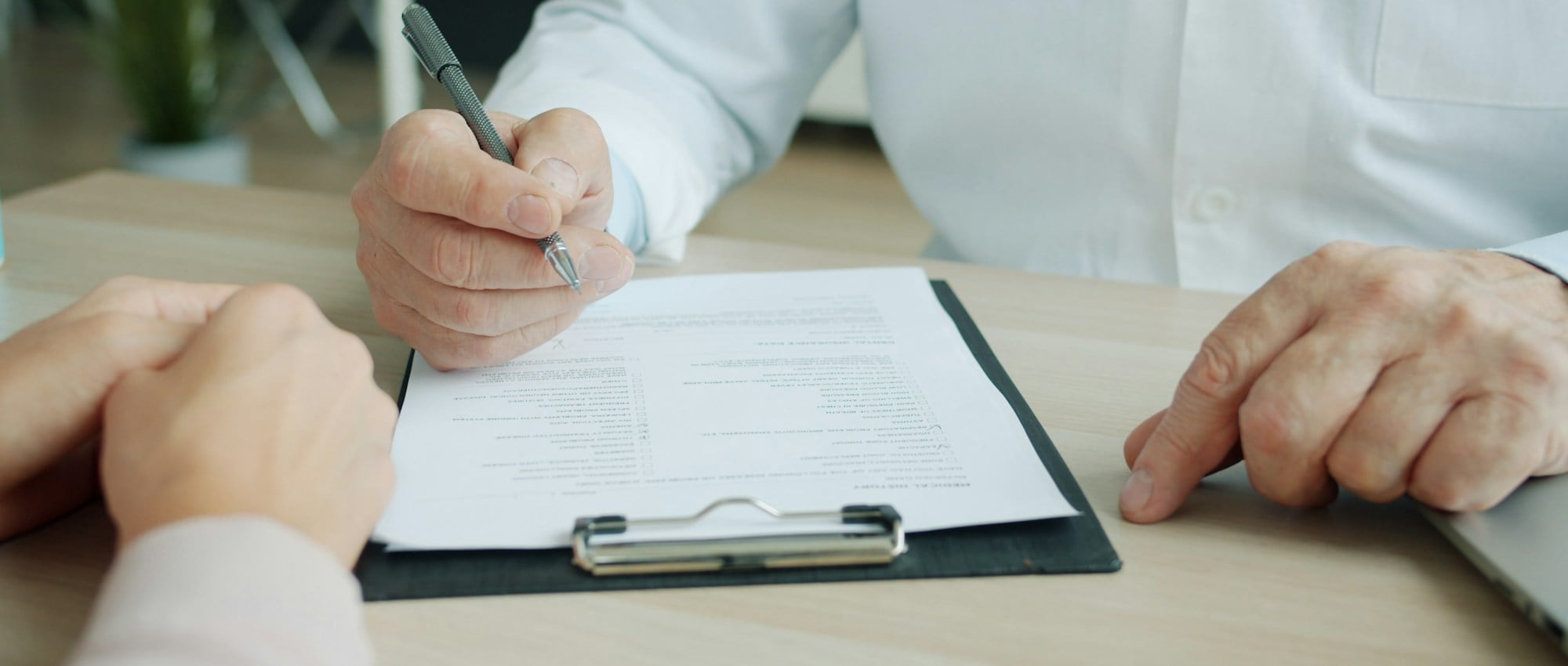 Physician taking notes on a clipboard across the desk from a patient during an assessment